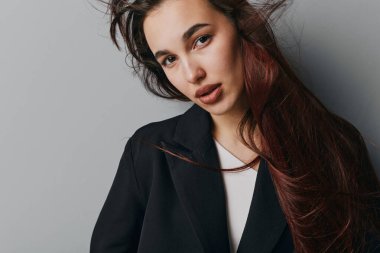 Siyah ceketli, beyaz gömlekli, dağınık saçlı ve makyajsız genç bir kadın portresi. Rahat bir ruh hali ve düz gri bir arkaplan ile kameraya bakıyor..