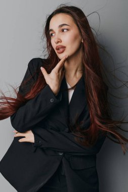 Portrait of a woman with flowing hair dressed in black jacket looking aside with thoughtful glance. Fashion model posing indoors with casual elegance and minimalistic style on gray backdrop.