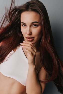 Portrait of young female with flowing brown hair wearing white sleeveless top. She looks directly at camera with soft smile and hand touching chin, studio shot with neutral backdrop.