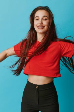 Happy young female with long hair in red shirt and black pants posing with arms out on blue backdrop. Joyful expression, casual fashion and energy for lifestyle and freedom.