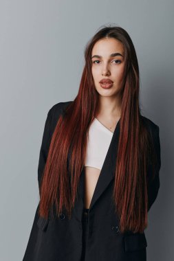 Portrait of confident young woman with long brown hair dressed in black jacket and white shirt posing in studio with neutral gray background and direct gaze.