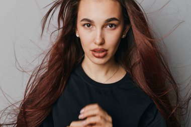 Sade gri duvara karşı poz veren dalgalı saçlı ve siyah bluzlu genç bir kadın. Kendine güvenen bir kadın modeli ve stüdyo resminde sıradan bir tarzı var..