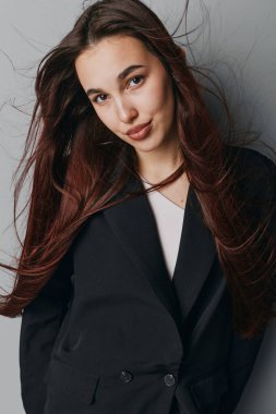 Portrait of young woman with brown hair in black blazer and white top. Female model posing indoors with calm expression, natural look, and casual hairstyle on neutral background.