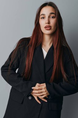 Siyah ceketli, uzun düz saçlı, gri arka planda duran genç bir kadının portresi. Katlanmış elleri ve sakin yüzüyle doğrudan kameraya bakan bir kadın model..