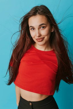 Confident young woman in red shirt and black pants standing in front of blue wall. Female model with long hair smiling with relaxed pose and casual fashion look.