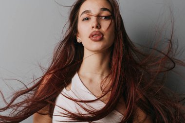 Portrait of young woman with brown hair blowing in wind. Female with natural look wearing white top standing in front of plain gray background.