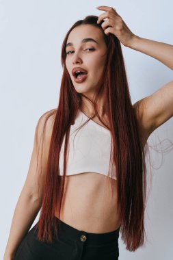 Portrait of confident woman with long brown hair touching head and smiling slightly wearing sleeveless white top and black pants on plain white background.
