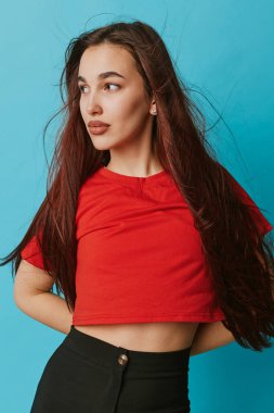 Portrait of young female with long hair in red top and black pants standing against blue backdrop. Stylish woman, casual outfit, natural look with earrings and confident expression for lifestyle