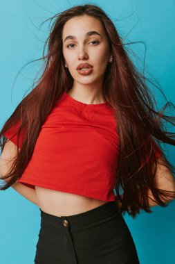Portrait of young woman with flowing hair in red shirt and black trousers standing on blue backdrop. Casual fashion look with minimal makeup and relaxed pose for lifestyle and elegance.