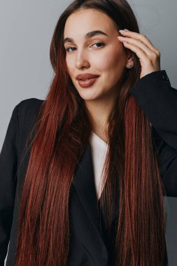 Uzun kızıl saçlı, ince makyajlı, siyah ceketli, beyaz tişörtlü, elinde gri arka plandaki kameraya bakan kendine güvenen kadın portresi.