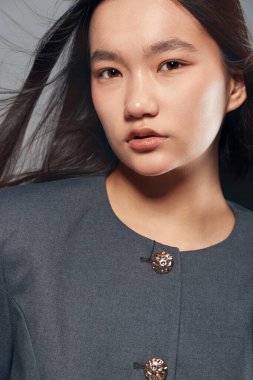 Stüdyoda yumuşak ışıklandırma ve nötr ruh haliyle metalik düğmeler içeren resmi gri ceket giyen pürüzsüz ciltli ve koyu renk saçlı genç kadın yüzü..