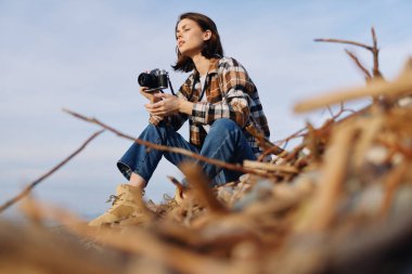 Kameralı bir kadın fotoğrafçının açık hava portresi. Etrafı sürüklenen odun ve plaj elementleriyle çevrili. Doğayı, ışığı ve sabit bir duruşta seyahat anlarını belgeliyor..