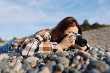 Kayalık bir sahilde odaklanmış bir kadınla açık hava fotoğrafçılık sahnesi, kamera yükselişte, günlük kıyafetler, dokuları keşfetmek, yaratıcılık için doğal ortamdaki ışık ve rüzgar..