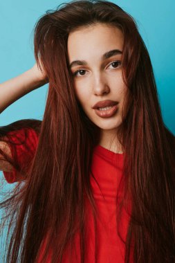 Portrait of young woman with long hair in red top standing near blue wall. Female model with relaxed pose touching hair and looking at camera with soft expression.