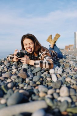 Çakıl taşlı plajda gülümseyen bir kadın fotoğrafçının gizli bir fotoğrafı, bir kamerayla manzara yakalamak, rahat moda tarzı ve gün ışığında sahil havası..