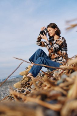 Kumlu kumullarda elinde kamerayla oturan kadın fotoğrafçı, okyanus manzarası ve yabani otları yakalıyor, açık hava çalışmalarını, odaklanmalarını ve yaratıcı hikaye anlatımlarını vurguluyor..