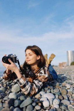 Çakıl taşlarının üzerinde uzanan kadın, el kamerası, günlük kıyafet, gevşemiş ruh hali, amatör ruh hali, doğa ilhamı, seyahat fotoğrafçılığı ile kıyı manzarasını yakalıyor..