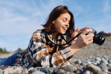 Çakıl taşlı bir plajda, güneşli mavi gökyüzüne ve açık ufka karşı küçük bir kamerayla çekim yaparken uzanan ve gülümseyen sıradan bir fotoğrafçı.
