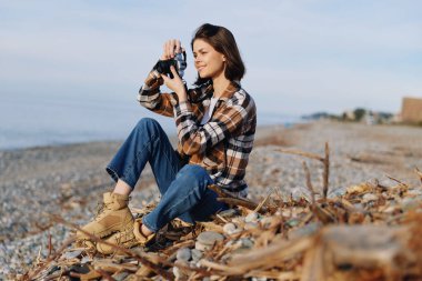 Sahildeki açık hava fotoğrafçısı kamera ve doğa sahnesi, ekose gömlek ve kotla günlük giysiler, sakin ufuk çizgisi ve huzurlu kıyı şeridi ile samimi bir ruh hali yakalıyor..