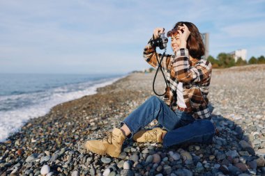 Kayalık bir sahilde bir kadın fotoğrafçı denizi kamerayla çerçeveler, günlük kot ve botlar giyer, açık deniz ışığında kompozisyon, ruh hali ve anlatım üzerine yoğunlaşır.