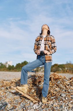 Kayalık bir sahilde, ekose gömlekli, kot pantolonlu ve botlu bir mankenle gündelik moda sahnesi. Açık mavi gökyüzü altında keşif, özgürlük ve rahatlama hissi veriyor.