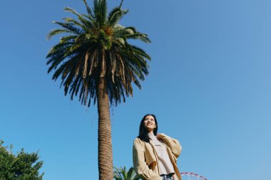 Avuç içi, ağaç, kadın, açık hava sahnesi, güneşli bir parkta güneşli bir gökyüzünün altında şık bir giysi ve eğlence sunan, özgürlük ve iyimserliği yakalayan bir manzara.