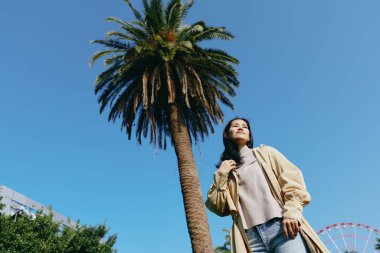 Açık hava sahnesinde bir kadın açık mavi bir gökyüzünün altında, uzun bir palmiye ağacının yanında rahat stil, güneş ışığı ve kaygısız yaz titreşimleri yayıyor.