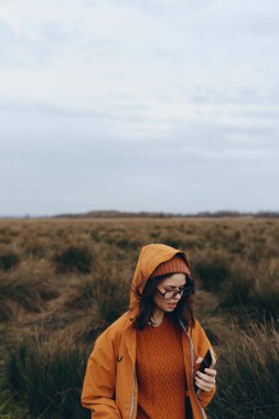 Açık alanda akıllı telefon kullanan turuncu ceketli ve gözlüklü genç kadın imajı. Sonbahar doğa arka planı bulutlu gökyüzü açık havada sakin ve düşünceli bir hava yaratıyor..