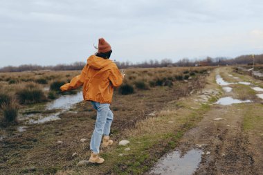 Kırsal alanda ıslak toprak bir yolda yürüyen kahverengi ceketli ve şapkalı bir kadının yaşam tarzı. Açık hava gündelik tarzı, sonbahar sezonu, doğa keşfi ve aktif kadın yaşam tarzı..