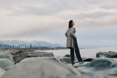 Paltolu genç bir kadın, bulutlu gökyüzünün altında, deniz kenarındaki taşlarda, dağlar ve uzaktaki palmiyeler üzerinde duruyor. Yansıma, yalnızlık ve doğal manzara teması.