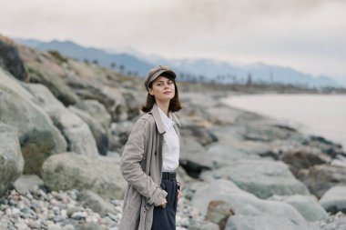 Paltolu ve şapkalı kadın, deniz ve dağlarla birlikte kayalık kıyılarda poz veriyor. Gündelik tarz, doğa ve açık hava yaşam tarzı kavramı yumuşak doğal ışıkla yakalanır..