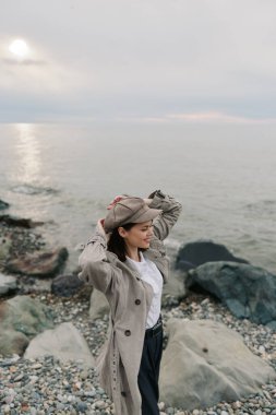 Şık kıyafetli bir kadın, gün batımında sakin okyanusun kıyısında kayalık sahilde şapka tamir ediyor. Sahne huzur, açık hava yaşam tarzı ve sakin bir sahil ortamında doğa ile bağlantıyı resmediyor..