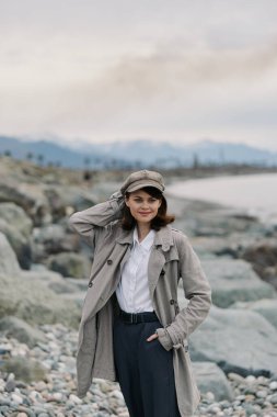 Smiling young woman in a hat and long coat poses on a rocky shoreline. Mountains and cloudy sky create a calm natural landscape with an autumn outdoor vibe and casual fashion.
