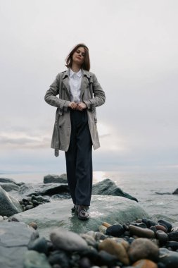 Gri bir ceket ve koyu renk pantolon giyen kendine güvenen genç bir kadın sakin deniz ve bulutlu gökyüzü ile kayalık kıyı şeridinde duruyor. Günlük kıyafet ve doğal çevre..