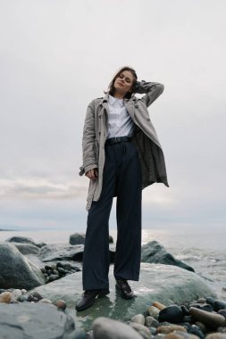 Uzun paltolu ve geniş pantolonlu, kayalık sahilde duran şık bir genç kadın. Suyun yanında, bulutlu gökyüzünün altında el üstünde poz veriyor. Sıradan bir stil ve doğa manzarası sergiliyor..