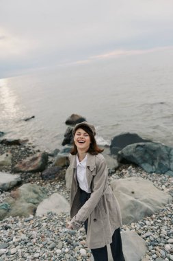 Palto ve şapka giyen mutlu genç kadın bulutlu gökyüzü ile sakin deniz kıyısında kayalık sahilde vakit geçiriyor. Gelişigüzel stil ve rahatlamış pozitif duygularla doğal açık hava ortamları..