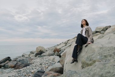 Kasvetli gökyüzünün altında sakin denizin yanında kayalık sahilde oturan sıradan giyinmiş bir kadın. Yalnızlık, rahatlama, doğa ve yumuşak doğal ışıkla huzurlu bir açık hava ortamı..