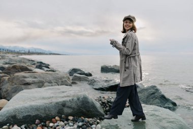 Paltolu ve bol pantolonlu gülümseyen kadın deniz kıyısında taşlarda yürüyor. Bulutlu gökyüzü ve sakin su açık hava yaşam tarzı ve doğa keşfi için sakin bir ortam hazırladı..