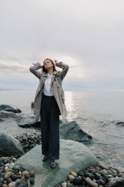 Paltolu ve geniş pantolonlu zarif bir kadın deniz kenarındaki taşın üzerinde duruyor. Sakin su ve bulutlu gökyüzü, o temiz havanın ve doğal kıyı manzarasının tadını çıkarırken sakin bir hava yaratıyor..