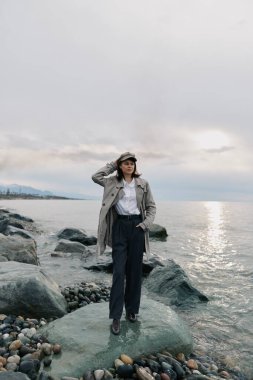 Paltolu ve şapkalı bir kadın okyanus arka planında kayalık sahilde poz veriyor. Bulutlu gökyüzü ve pürüzsüz su doğal kıyı manzarasını ve günlük moda tarzını ön plana çıkarıyor.