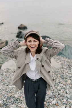 Smiling young woman wearing hat and stylish jacket enjoys day on rocky shore by water. Candid portrait with natural background, casual outfit, and positive mood outdoors.