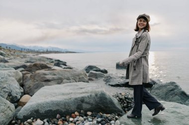 Şapkası ve paltosu olan gülümseyen kadın denizin kıyısındaki kayalık sahilde duruyor. Bulutlu gökyüzü ve çakıl taşları kasvetli bir sonbahar sahnesi yaratır. Doğa, seyahat ve açık hava modası kavramı.