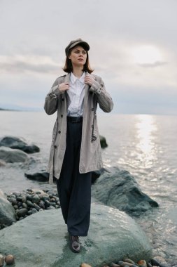 Kareli ceket ve şapka giymiş kadın model gün batımında su kenarında taşların üzerinde poz veriyor. Yumuşak doğal ışıkta rahat stil ve karamsar kıyı manzaralı açık hava modası..