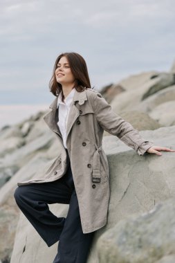 Gri ceketli ve siyah pantolonlu gülümseyen genç kadın kıyıya yakın kayalarda oturuyor. Bulutlu gökyüzü ve doğal taş manzarası, sahneye sakin ve huzurlu bir hava katar..