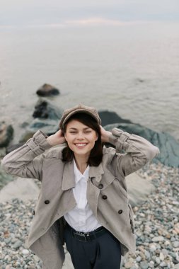 Happy young woman wearing a hat and textured coat stands on a pebble beach near calm water, smiling with hands behind head. Concept of casual fashion, autumn style, and outdoor relaxation.