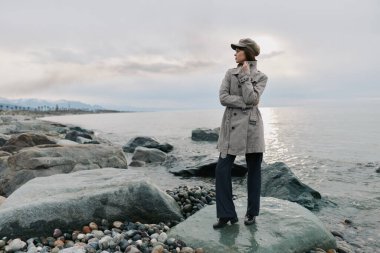 Gri paltolu ve siyah pantolonlu şık bir kadın bulutlu gökyüzünün altında sakin denizin yanındaki kayalık sahilde duruyor. Sonbahar stili, doğa ve barışçıl doğa kavramı..