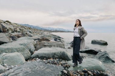 Sıradan giyinmiş bir kadın bulutlu gökyüzünün altında sakin okyanus kıyısında kayalık kıyılarda yürüyor. Güvenli bir şekilde poz veriyor ve sakin bir günde temiz hava ve doğal ortamın tadını çıkarıyor..