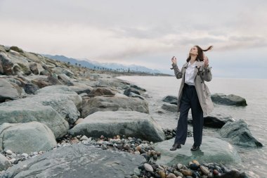 Paltolu ve siyah pantolonlu kadın bulutlu gökyüzünün altında sakin okyanusun kenarında büyük kayaların üzerinde duruyor. Özgürlük kavramı, doğa, açık hava yaşam tarzı ve su ve taşların yanında dikkatli bir an..