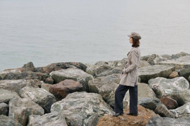 Kareli paltolu ve şapkalı kadının okyanus kıyısındaki büyük kayalıklarda duruşu. Bulutlu gökyüzü ve yumuşak ışıkla sakin bir atmosfer sakin, yansımalı ve açık havada keşifleri çağrıştırıyor..