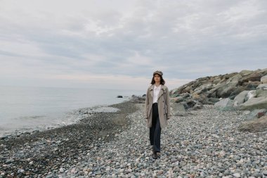 Paltolu ve şapkalı genç bir kadın bulutlu gökyüzünün altında sakin deniz kıyısında kayalık kıyılarda yürüyor. Kaya ve çakıl taşlı karamsar doğa sahnesi. Yalnızlık, sonbahar yürüyüşü ve huzurlu bir açık hava yaşam tarzı..